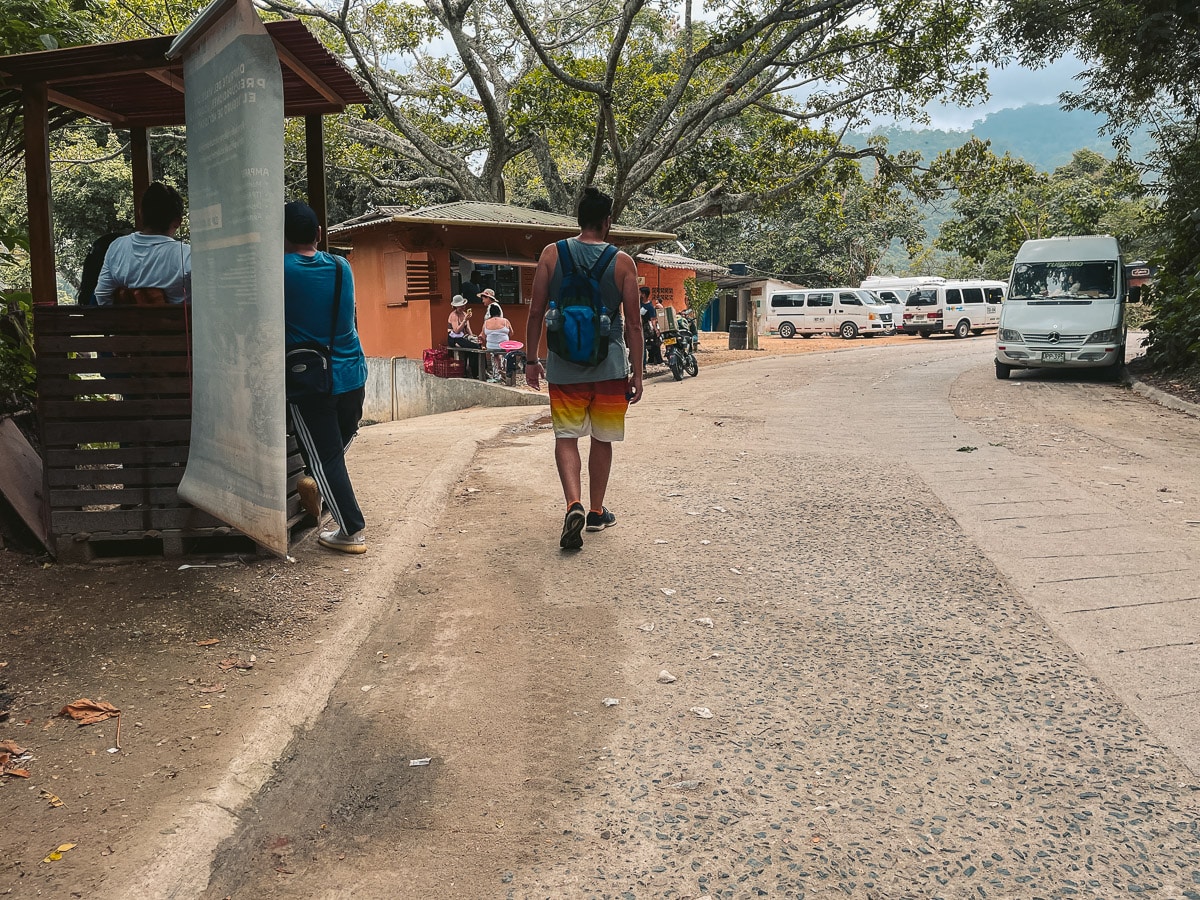 A person wearing a blue backpack and colorful shorts walks down a rustic, tree-lined road toward a small building where people are seated at the Pozo Azul entrance. Multiple white vans are parked along the road under a cloudy sky.